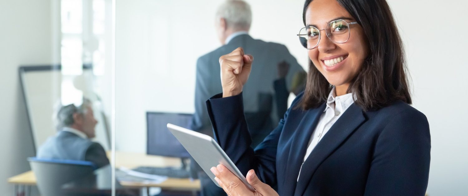 happy-female-professional-in-glasses-and-suit-holding-tablet-and-making-winner-gesture-while-two-businessmen-working-behind-glass-wall-copy-space-communication-concept (1)